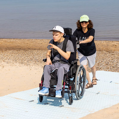 Person in wheelchair on beach mat. Another person is pushing.