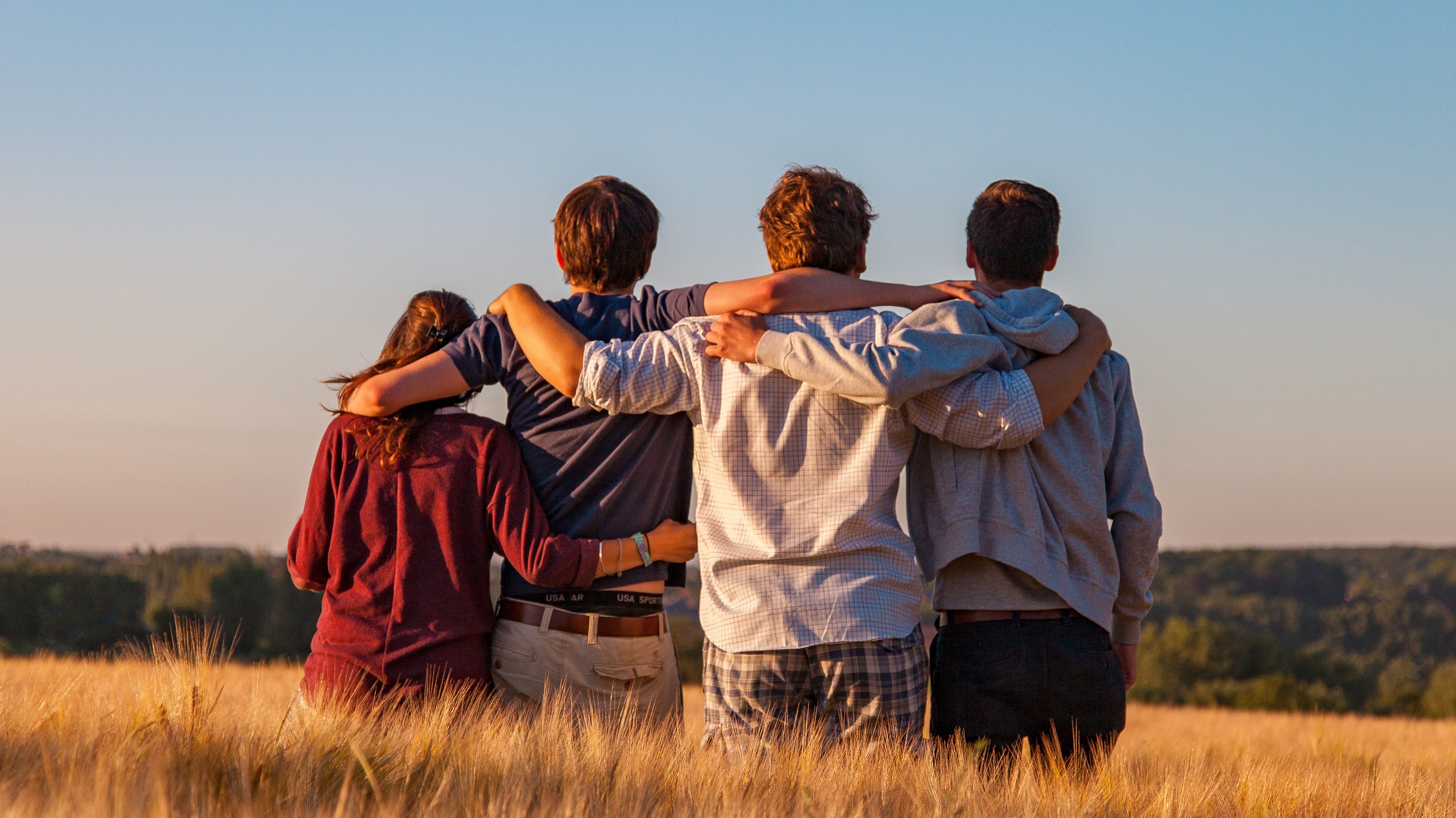 Four youths link arms around each other's backs while standing in a wheat field looking away from the camera towards woods.