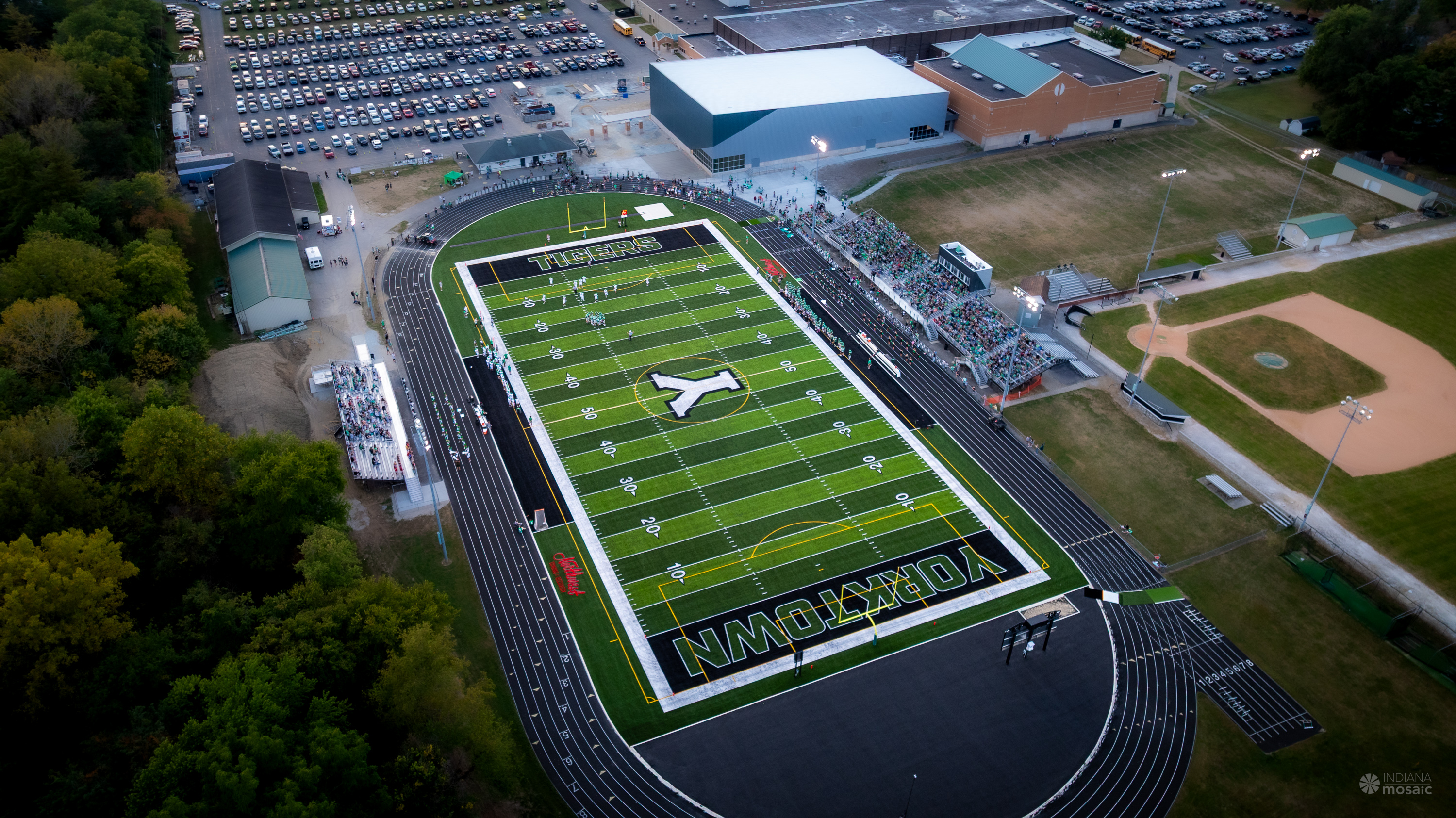 Aerial view of Yorktown High School's athletic complex during a football game at dusk. The image shows the Tigers football stadium with a lit artificial turf field surrounded by a running track, grandstands filled with spectators on both sides, the high school buildings beyond the stadium, adjacent baseball fields, and expansive parking areas, highlighting the campus's modern athletic facilities.