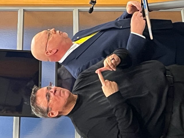 Man in all black clothing stands next to a man in a blue suit with yellow tie.
