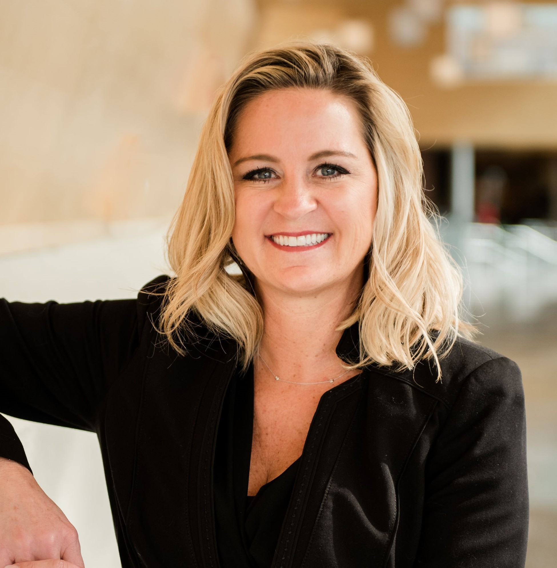 A person with light-colored, shoulder‑length hair is shown in a close-up portrait, wearing a dark jacket and smiling while positioned in an indoor setting with warm, blurred background elements.