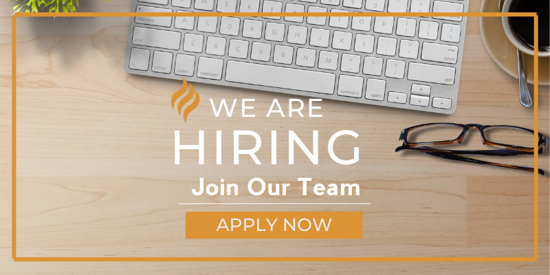 A keyboard, cup of coffee, and pair of glasses lay on a wooden table, and the words "WE ARE HIRING. Join Our Team. APPLY NOW."