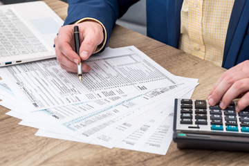 A close-up of hands working on tax forms. One hand is writing with a pen, and the other is typing on a calculator.