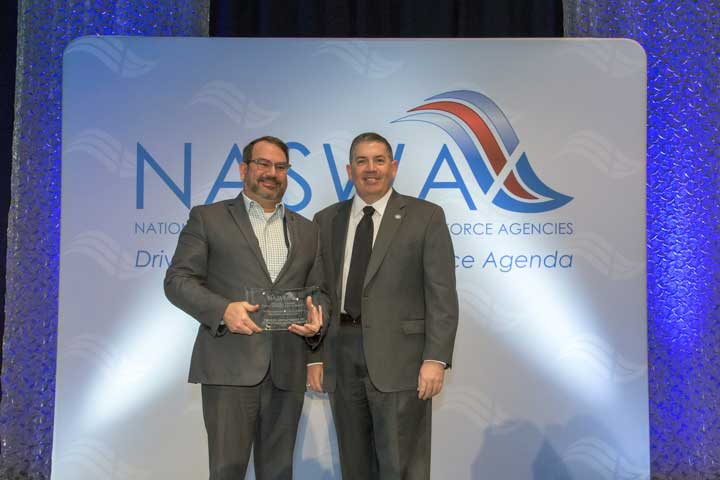 Picture of DWD Staff Holding NASWA Award in Front of NASWA Banner on Stage