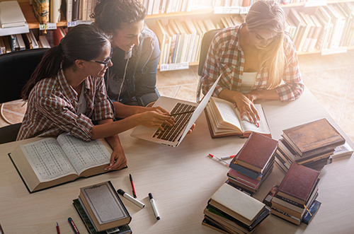 three students study books at a table in a library