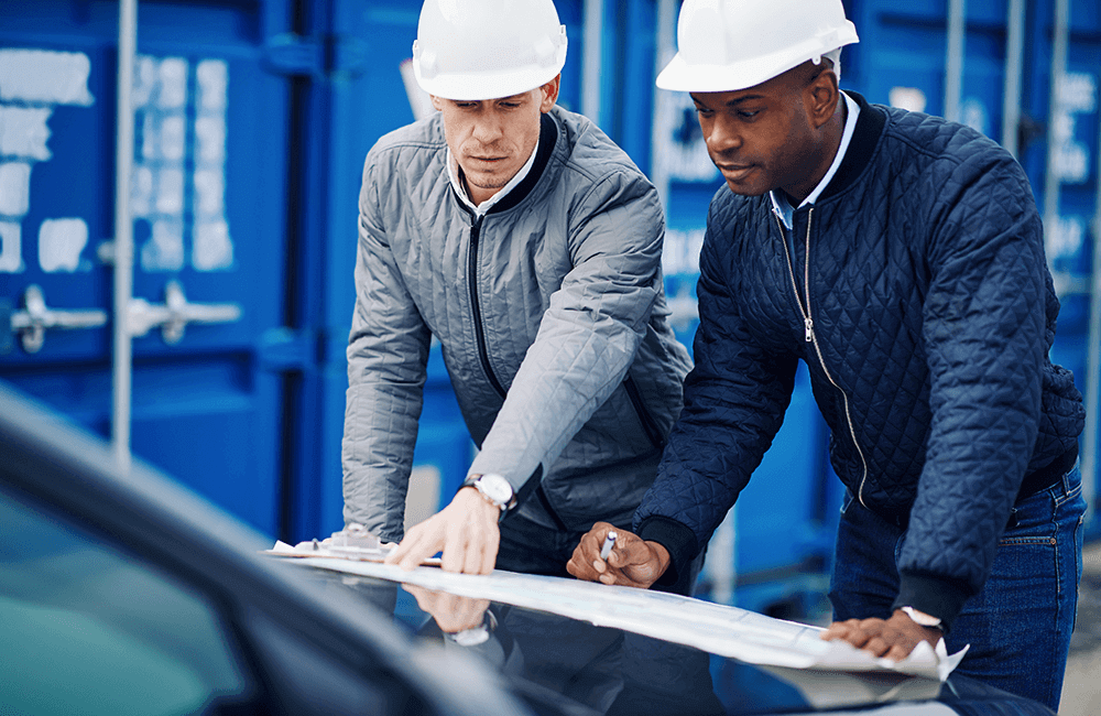 Two Male Construction Workers Review Blue Prints on the Hood of a Vehicle in Front of Shipping Crates