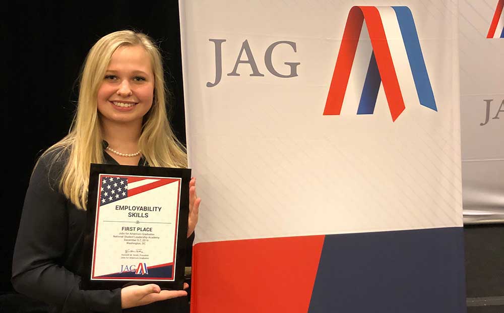 JAG Student Winner Holding Plaque for First Place in Employability Skills Competition Standing in Front of JAG Poster