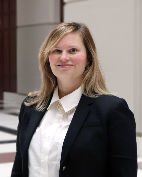 Portrait of Allison Wymer, a white person with shoulder-length blond hair, wearing a white blouse and black jacket, with a pearl necklace. She is smiling and standing beneath a bright atrium, with tile floors and tall white pillars behind her.