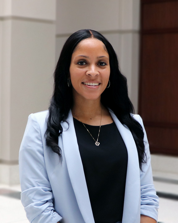 Portrait of Desirea Island, a black woman with long dark hair, wearing a powder blue blazer and a black blouse, with a small silver pendant necklace and hoop earrings. She is smiling and standing beneath a bright atrium, with tile floors and white walls and pillars behind her. 