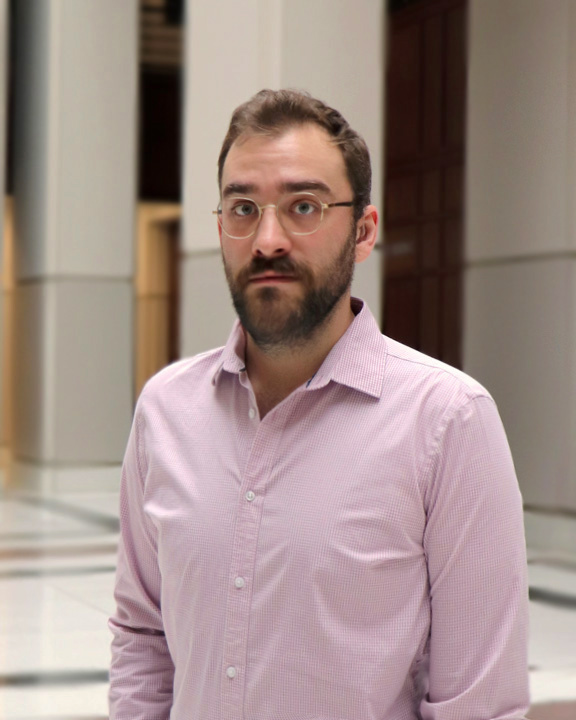 Portrait of Hayden McGough, a white man with short dark blond hair and a scruffy beard, wearing gold wire-frame eyeglasses, a hearing aid, and a pale pink dress shirt with its collar open. He has a calm, patient expression while standing beneath a bright atrium, with tile floors and tall white pillars behind him.