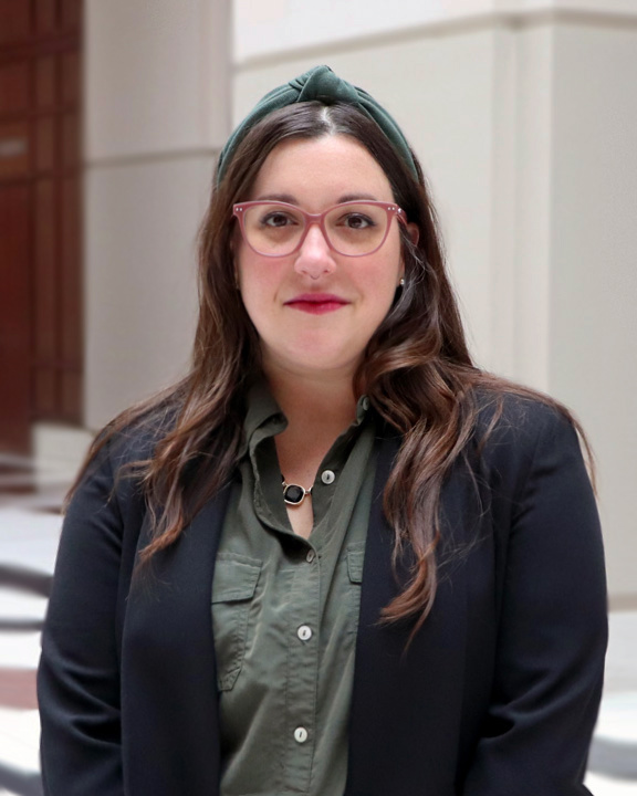 Portrait of Kate Barrow, a white woman with long brown hair, wearing pink eyeglasses, an olive green blouse and headband, and a black jacket, with earrings, a septum nose ring, and a black-jewel necklace. She is smiling gently and standing beneath a bright atrium, with tile floors and white walls behind her. 