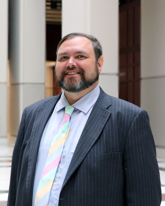 Portrait of Ryan Revell, a white man with dark hair combed to the side and a salt-and-pepper beard, wearing a pinstripe blazer, a light blue dress shirt, and a pink, yellow, and green striped necktie. He is smiling and standing beneath a bright atrium, with tile floors and tall white pillars behind him.