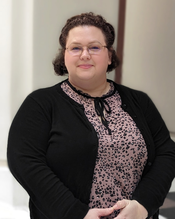 Portrait of Robin Martinez, a woman with dark curly hair pinned back, wearing wire-frame eyeglasses, a mauve animal-print blouse, and a black sweater. She is smiling and standing beneath a bright atrium, with white walls behind her.