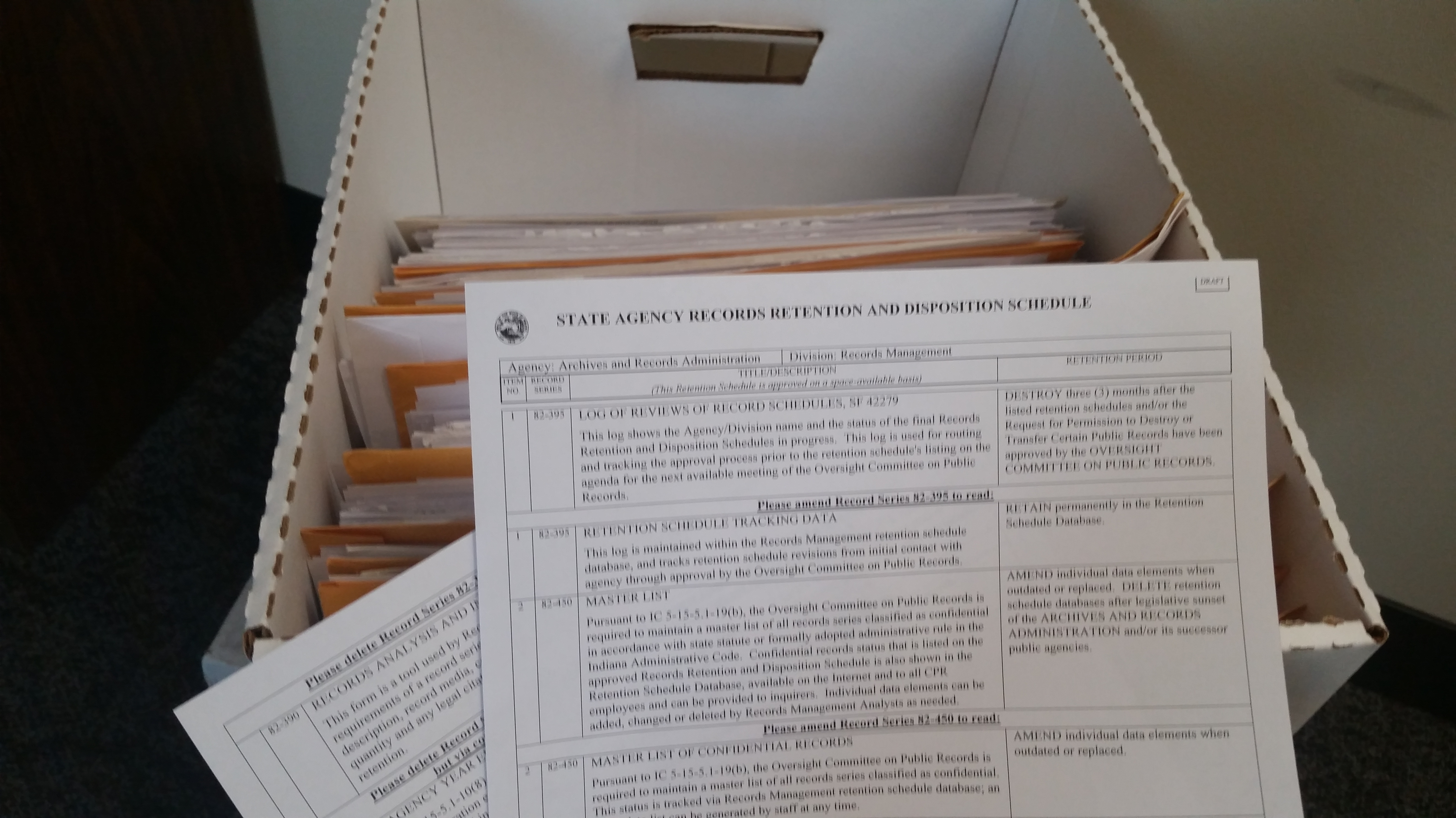 Photo of a hand holding a records retention schedule, in front of and above an open filing cabinet drawer
