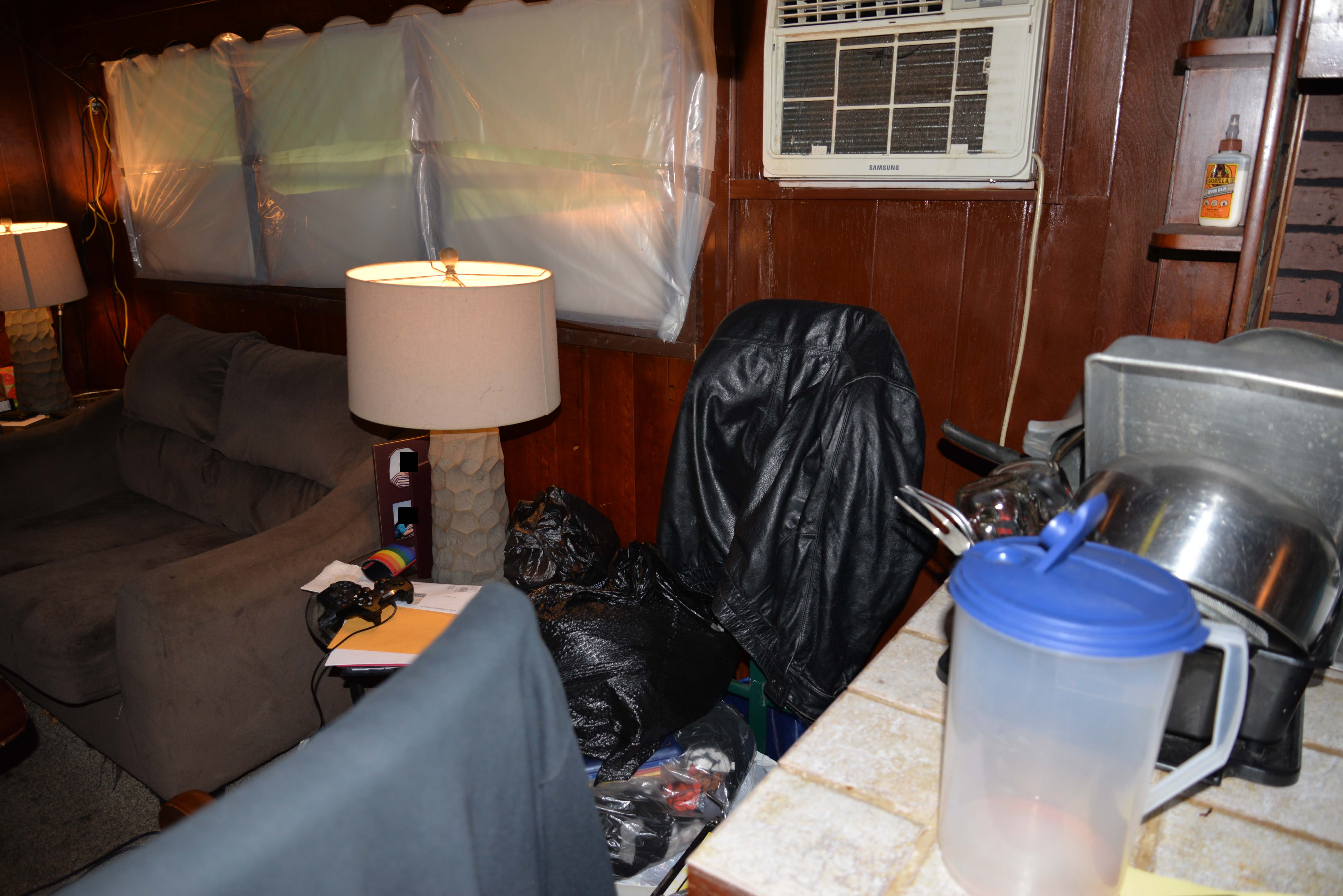 Aaron&rsquo;s living room has dark wood paneled walls, and an opaque white tarp covers the three windows directly above a gray couch. An in-wall air conditioner is mounted to the right of the windows. There is an end table on each side of the couch, both with a stone lamp. Multiple wires hang from the wall behind the left end table. The surface of the right end table is covered in papers, a PlayStation controller, rainbow beer koozie, and partially obscured photos. Farther to the right, two black garbage bags are resting on a clear bag stuffed with fabric. A black leather jacket hangs between the air conditioner and garbage bags. A tiled counter with an empty pitcher, pot, pans, and other utensils is further right. Small shelves, one of which contains Gorilla wood glue, are above. 
