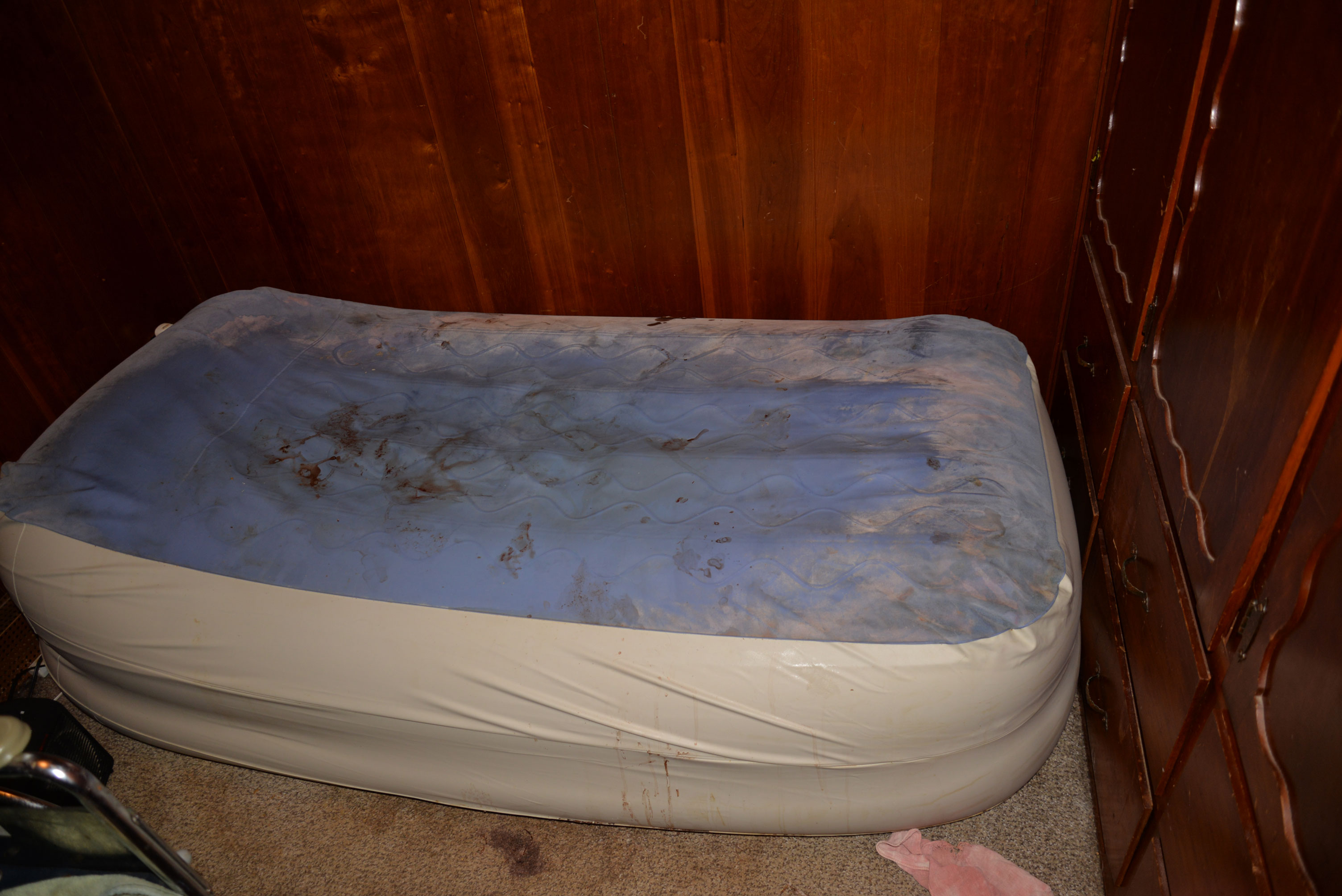 Aaron’s small mattress fills the width of a dark brown wood-paneled room. The mattress has no sheets, and its plastic cover has thick dust around the perimeter. Dried blood stains are smeared in the middle of the mattress. The mattress lays directly atop the stained, neutral-toned carpet. 