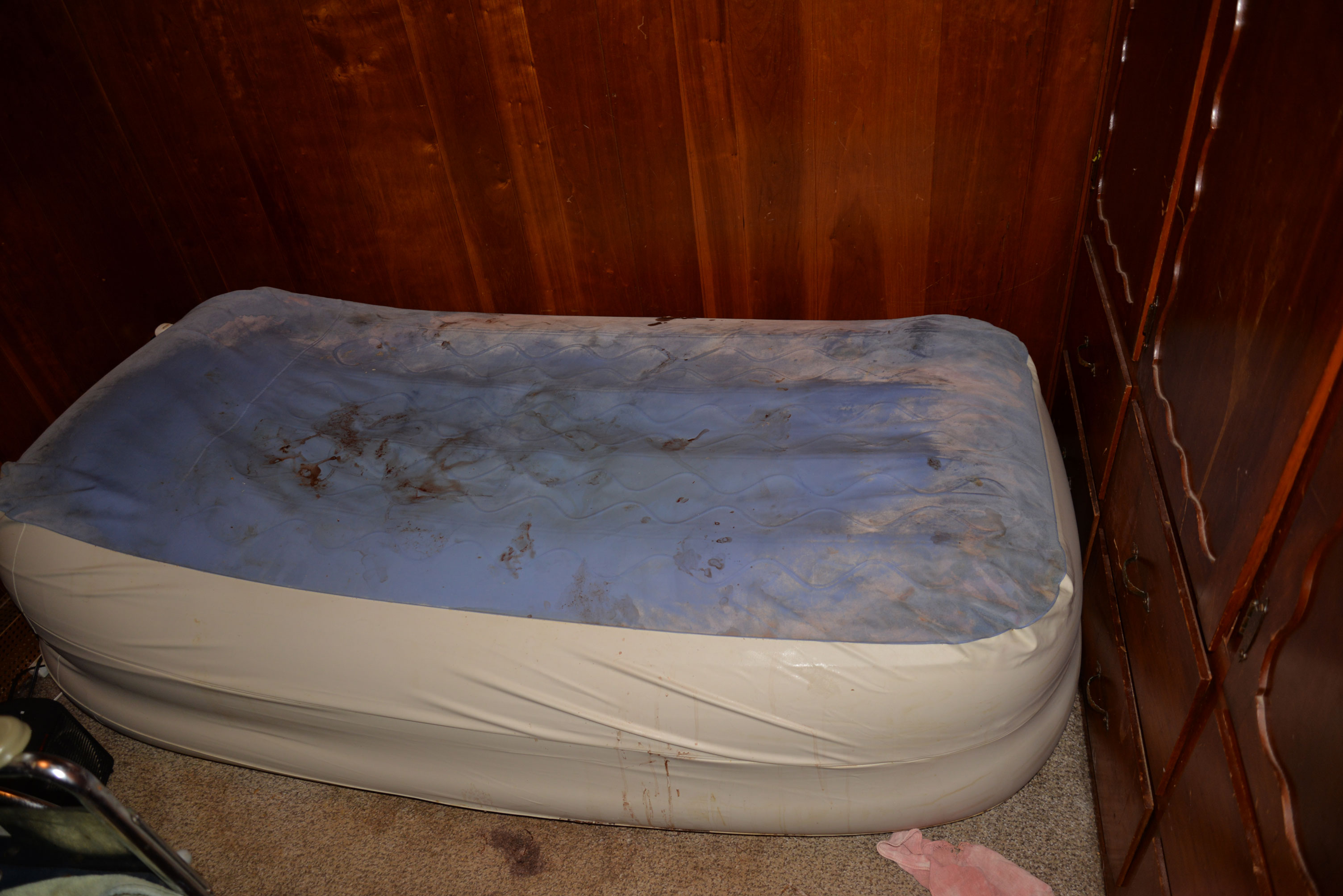 Aaron&rsquo;s small mattress fills the width of a dark brown wood-paneled room. The mattress has no sheets, and its plastic cover has thick dust around the perimeter. Dried blood stains are smeared in the middle of the mattress. The mattress lays directly atop the stained, neutral-toned carpet. 