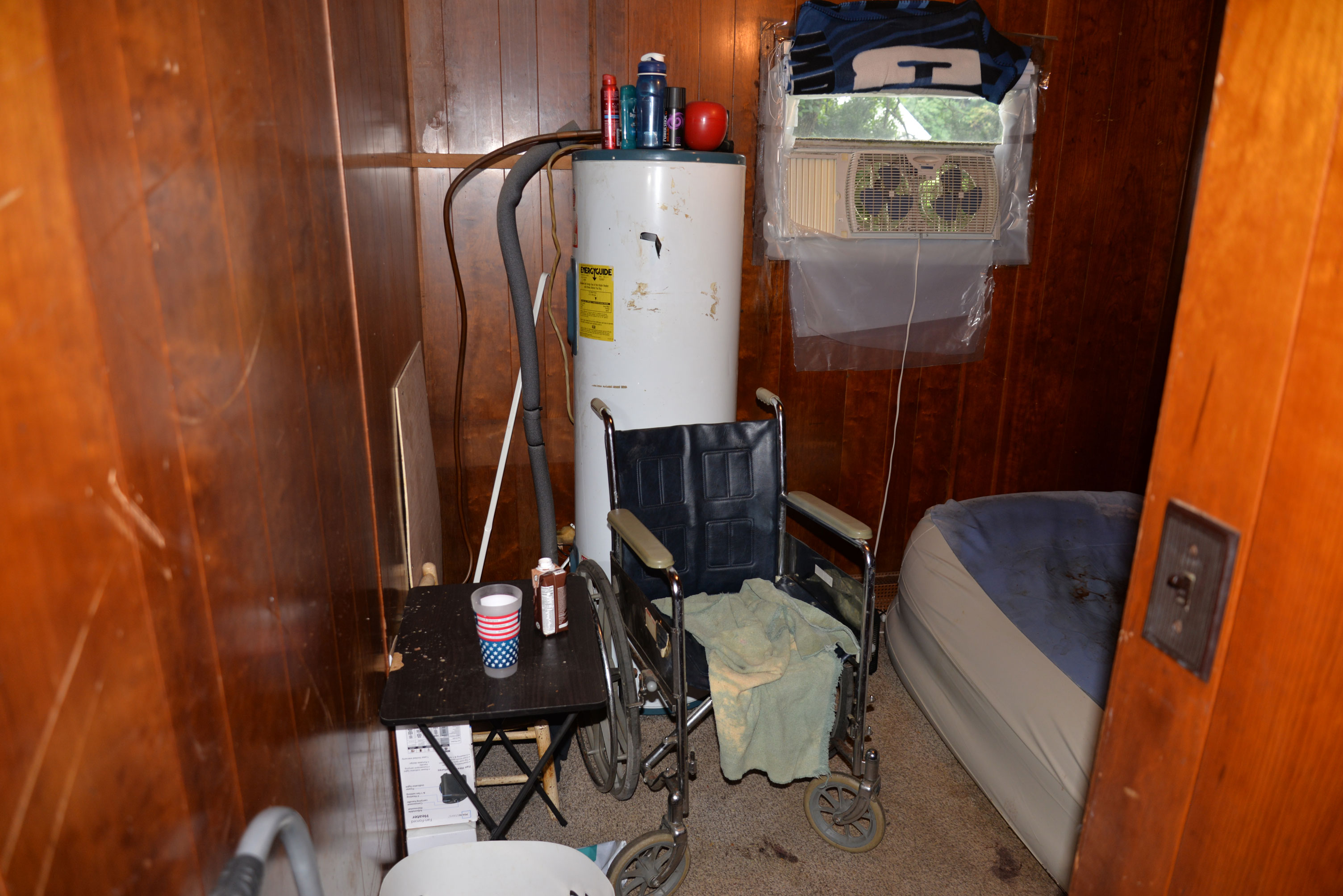 This picture shows side of Aaron&rsquo;s room across from his mattress, the corner of which can be seen on the right side of the photo. Scratches are visible in the dark brown wood wall paneling. The trailer&rsquo;s water heater stands in the back left corner. Between the mattress and the heater is a small window with a double boxed fan inside. The window is surrounded in plastic, which hangs several inches below the sill. Plastic surrounds the window and is hanging down from the window for several inches. A generic folding wheelchair sits in front of the water heater, with a frayed gray towel hanging from the seat. A small, dark, scratched TV tray table is opened near the wheelchair. Atop it are a cup with an upside down US flag and an individual serving-sized cardboard container of chocolate milk. 