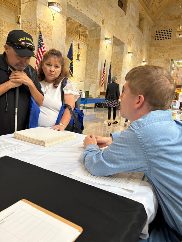 Man sits behind a table talking with a man and woman.