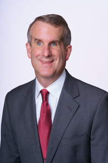 Man in a gray suit, white shirt, and red tie, who has light skin, blue eyes, and dark hair.