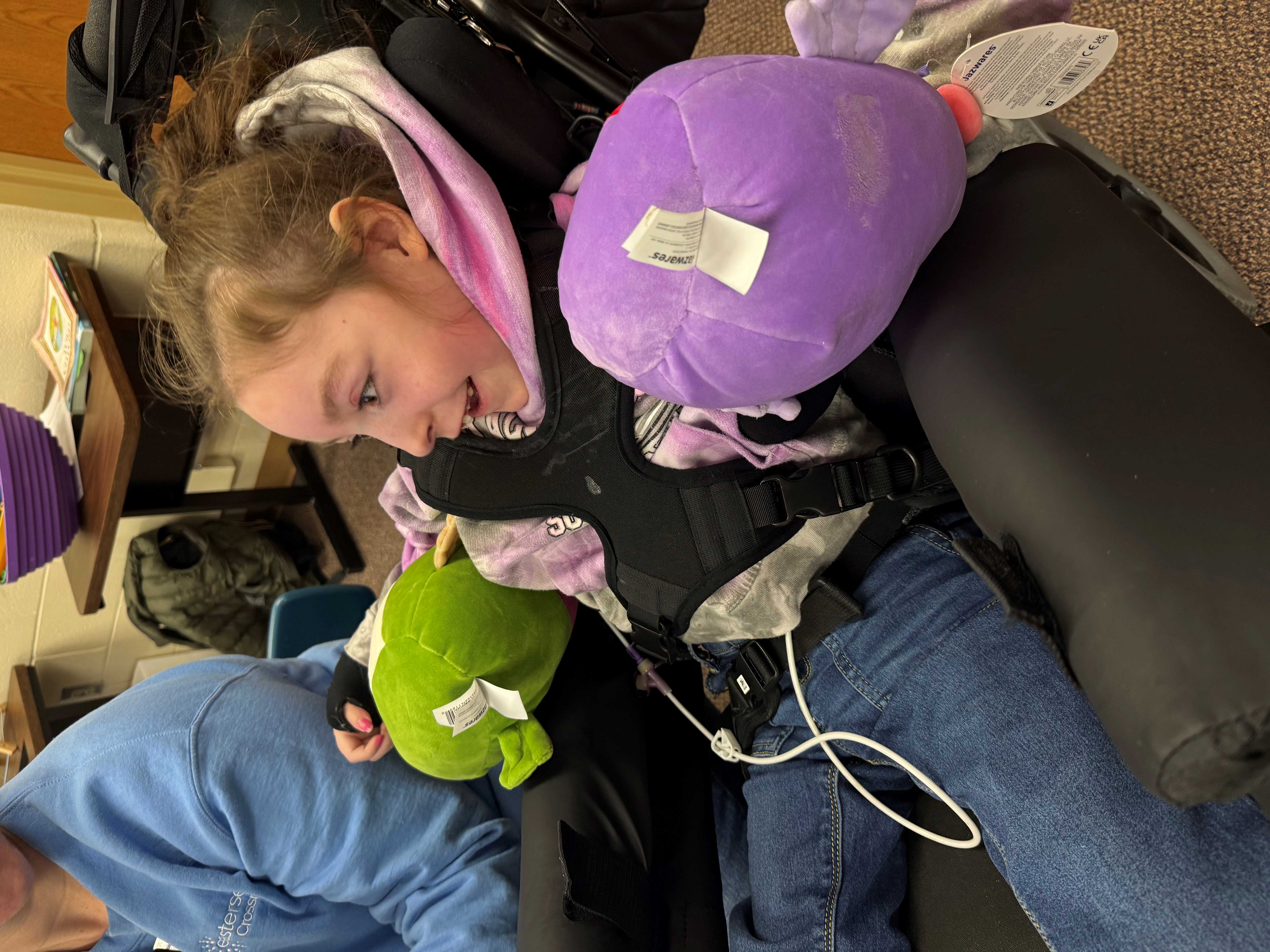 Sophia, a young girl with brown hair, wears jeans and a pink hoodie. She sits in a wheelchair with stuffed toys under her arms, smiling as she uses a communication device with support from a staff member in a blue sweatshirt. They are in her classroom, engaging with her offscreen teacher.