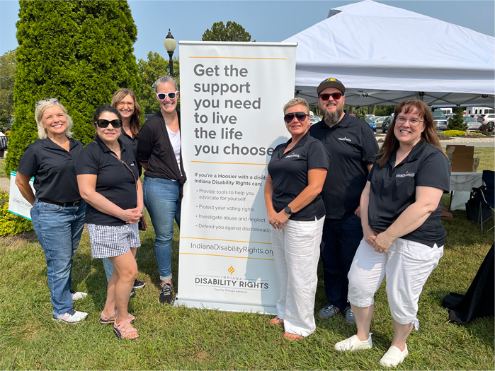 At an outdoor event, seven IDR staff members pose around a pull up sign that says "Get the support you need to live the life you choose."