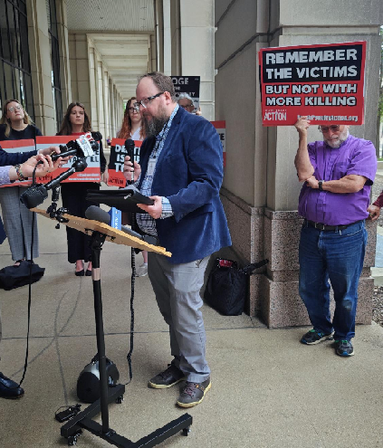 Sam Adams, a man in a suit, speaks at a microphone, surrounded by the media and community members, some of whom are holding signs.