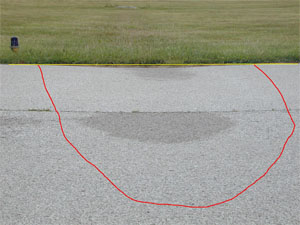 Photo showing a large-severity depression on the edge of an asphalt pavement.           A stringline has been stretched across the depression to illustrate the depth, and some water staining on the pavement surface is noticeable at the center of the depression.           A red line has also been drawn on the photo to outline the area with the depression.