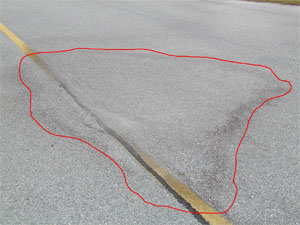 Photo showing a medium-severity depression on the pavement. The area of the           depression is noticeable due to water staining on the pavement. A red line has also been drawn on the photo to outline the area with the depression.