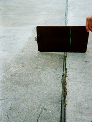 Close-up photo showing a transverse joint with a slight difference in elevation across the joint.           A clipboard is placed across the joint in the photo to illustrate that there is a height differential between the two slabs.