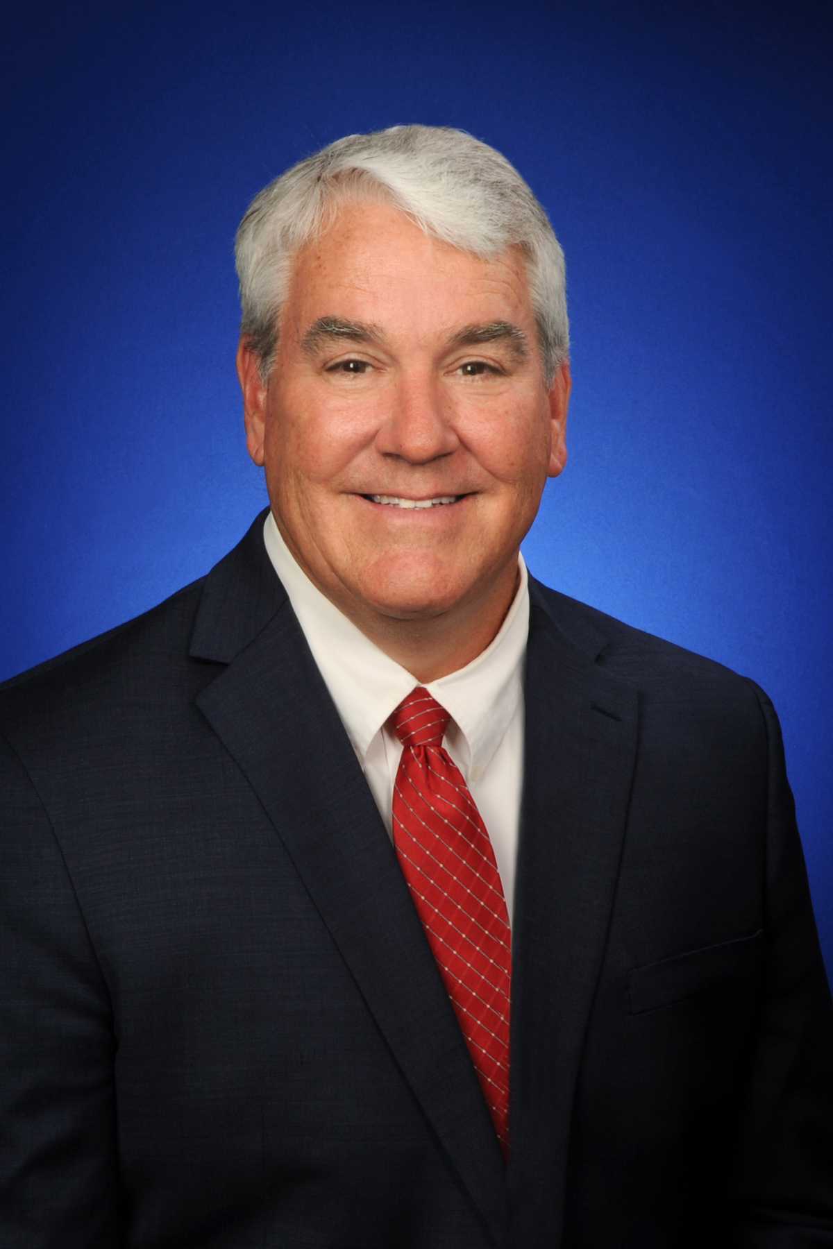 Chairman Zay is wearing a blue suit with a white shirt and red tie next to the Indiana state flag against a gray background.