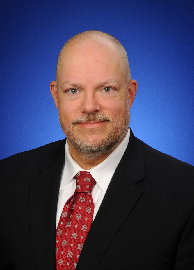 <img alt="A photo of IURC Commissioner Anthony Swinger dressed in a brown suit jacket with a black shirt and red tie, against a light brown brick background." />