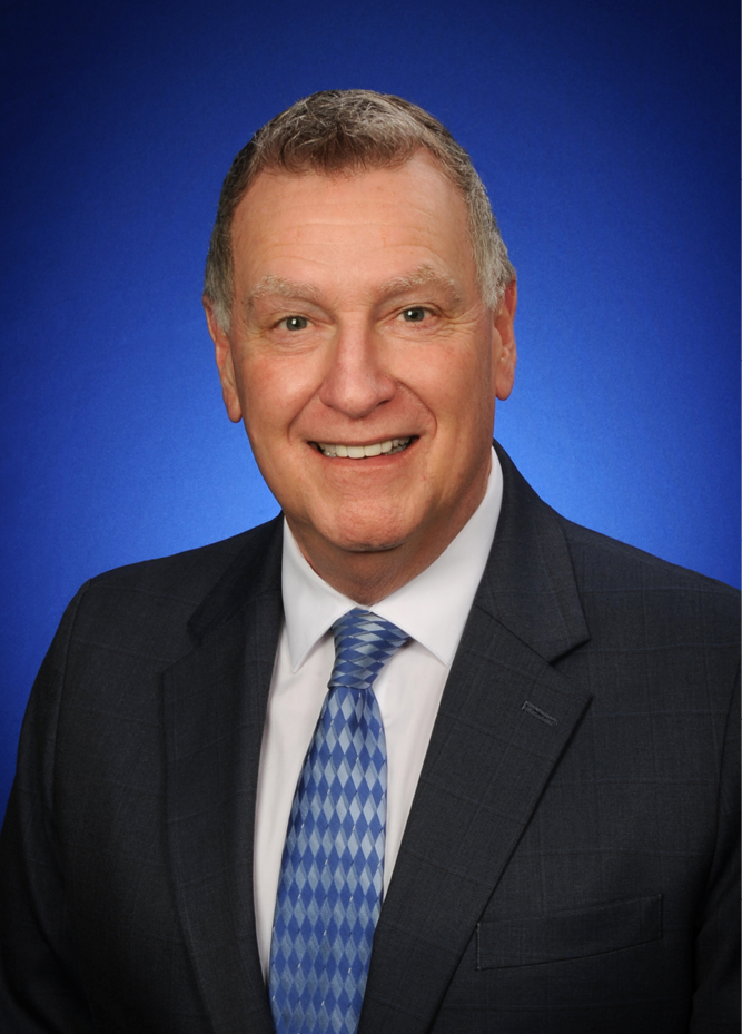 <img alt="A photo of IURC Commissioner Bob Deig, dressed in a pink suit, against a dark blue background." />