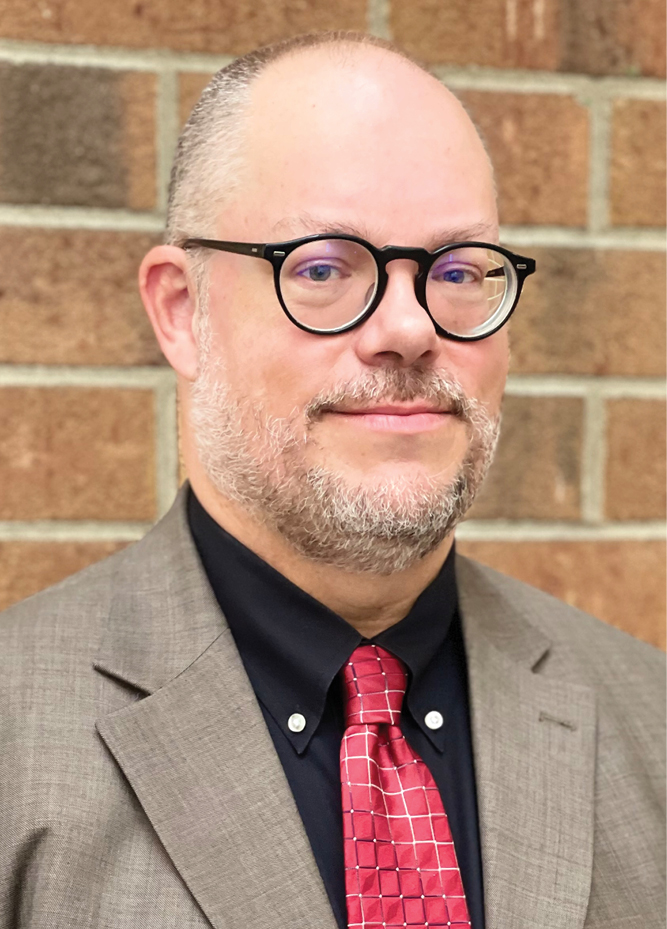 <img alt="A photo of IURC Commissioner Anthony Swinger dressed in a brown suit jacket with a black shirt and red tie, against a light brown brick background." />