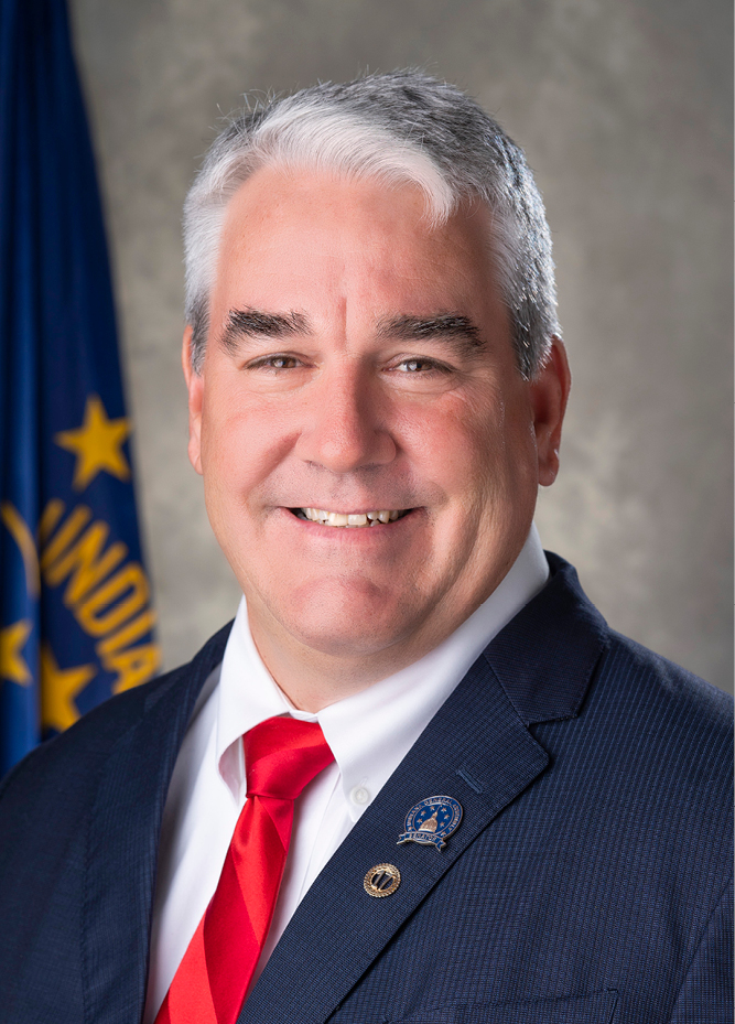 <img alt="A photo of IURC Chairman Andy Zay, dressed in a blue suit, standing by the Indiana state flag against a gray background." />
