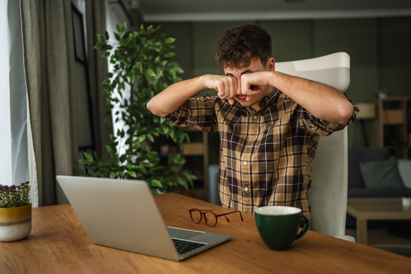 Young man sitting in front of a laptop, rubbing his eyes.